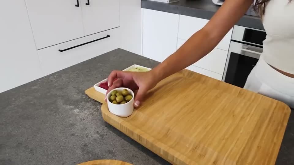 Hands arranging charcuterie and bowls on the bamboo cheese board in a kitchen